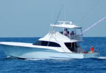Luxury sport fishing yacht on open ocean with fishing rods; white boat with flybridge and crew member; calm blue sea background.