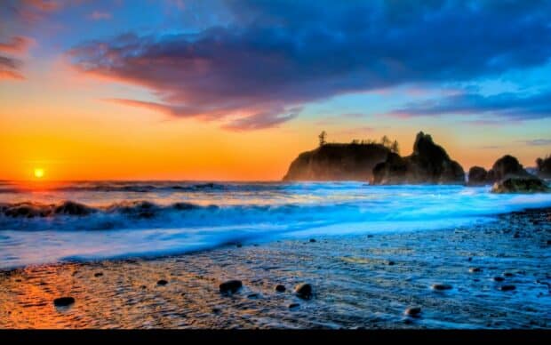 Sunset over rocky coastline with waves crashing, vibrant orange and blue sky, scenic Pacific Ocean landscape, evening seascape, tranquil beach scene, colorful sky, nature photography.