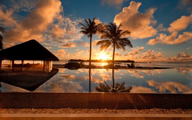 Tropical sunset over a pool with palm trees and a thatched hut, reflecting vibrant orange and blue sky, creating a serene island paradise scene.