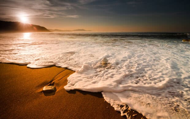 Sunset over sandy beach with waves crashing and foam, scenic ocean view at dusk, golden light, calm sea, coastal landscape, twilight, serene shoreline.