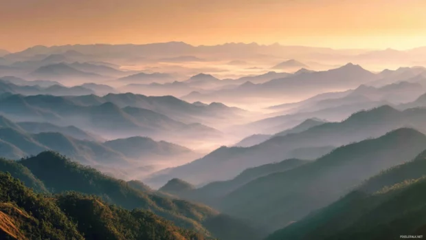 Widescreen view of a serene mountain range at sunrise with layers of mist drifting through the valleys.