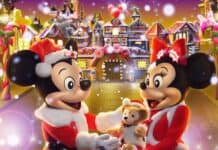 Festive Disney Christmas characters Mickey and Minnie Mouse in holiday costumes, smiling holding a teddy bear, with a colorful gingerbread house and Christmas decorations in the background.
