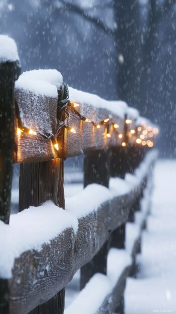 Snow covered wooden fence with Christmas lights glowing warmly in falling snow