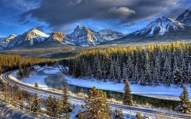 Snow covered pine trees and mountains in a winter wonderland landscape with a winding river and railway tracks
