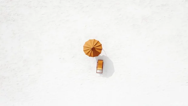 A minimalist design featuring a single beach chair and umbrella on a white sand background.