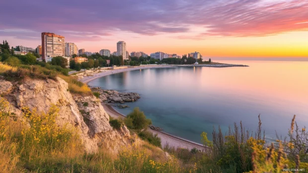 A panoramic view of a coastal skyline with modern buildings overlooking a calm sea, under a pastel pink evening sky.