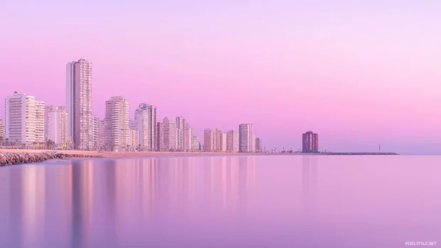 A panoramic view of a coastal skyline with modern buildings overlooking a calm sea, under a pastel pink evening sky.