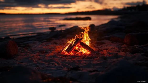 Bonfire burning on beach as sun sets, glowing on sand and rocks.