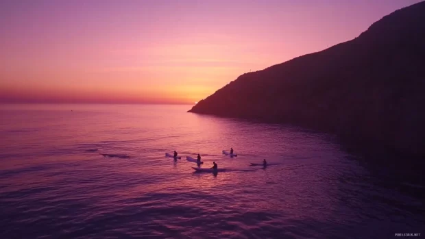 Surfers paddling out to catch the last waves at sunset, with a calm ocean and colorful sky.