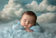 A baby boy wearing a soft blue onesie, lying on a cozy blanket with fluffy clouds in the background.