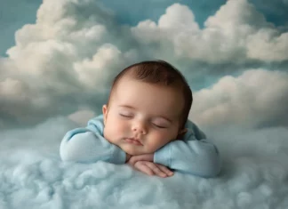 A baby boy wearing a soft blue onesie, lying on a cozy blanket with fluffy clouds in the background.