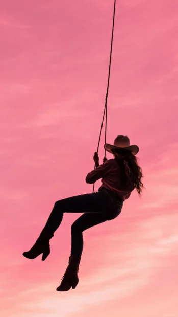 Cowgirl swinging on a rope in front of a pink sky.