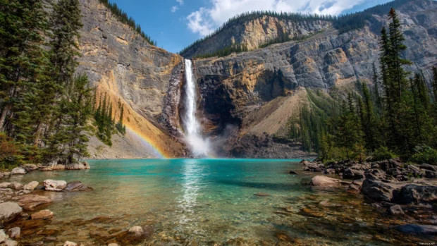 A colorful rainbow rises behind the Waterfall as it pours into a shimmering turquoise lake.