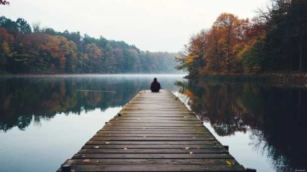 An old wooden dock stretching into a peaceful lake with someone fishing at the end.