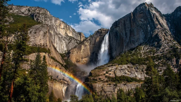 Rainbow falls HD wallpaper presenting majestic waterfall with strong rainbow arch and scattered mist over alpine mountain backdrop.