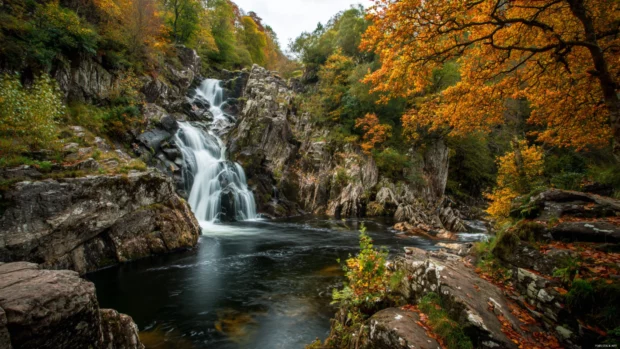 Waterfall tumbling over ancient stone formations surrounded by thick autumn foliage in crisp 4K clarity.