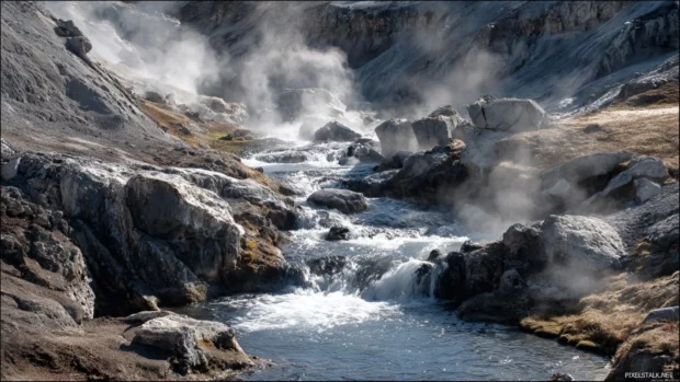 Waterfall tumbling over volcanic rocks with patches of steam rising in the cool air.