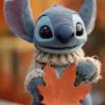 A close up of Stitch holding a large maple leaf, with Fall colors softly blurring in the background.