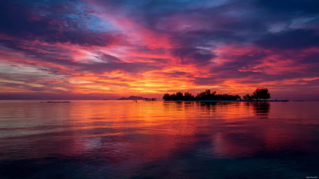 Ocean sunset wallpaper portraying fiery sky reflected on calm water with silhouette of distant island.