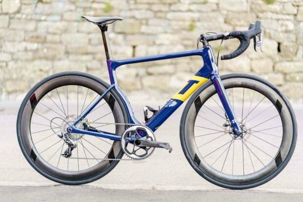 A blue and yellow 3T bike with high performance components is shown standing on a paved surface in front of a stone wall, HD Desktop Wallpaper