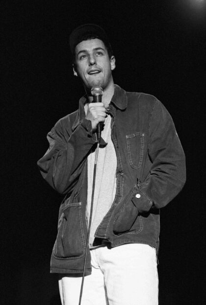 Adam Sandler is holding a microphone and wearing a jacket while performing on stage, 2K Phone Wallpaper