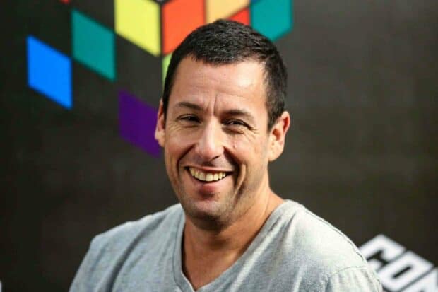 Adam Sandler is smiling warmly wearing a gray shirt with colorful geometric shapes in the background, 2K Desktop Wallpaper