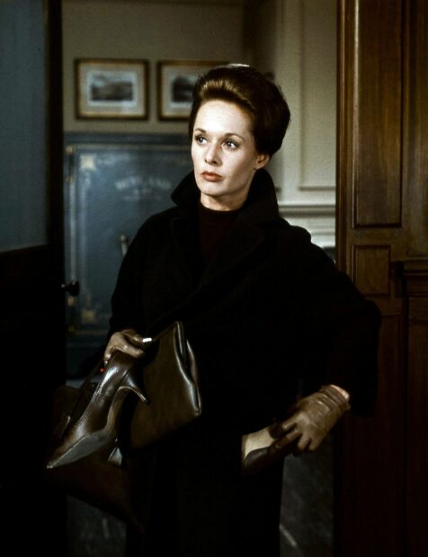 A woman in a black coat holding shoes and a handbag in a dramatic scene from an Alfred Hitchcock film, 2K PC Wallpaper