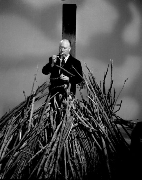 Alfred Hitchcock is lighting a cigarette while standing tied to a post surrounded by a pile of firewood, 2K Mobile Wallpaper