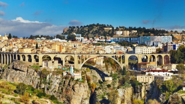 A historic bridge spans a deep gorge with buildings and hills in the background showcasing Algeria city life, 2K Desktop Wallpaper