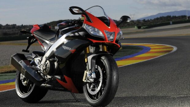 A red and black Aprilia RSV4 motorcycle parked on a curving race track under a clear sky, 2K Desktop Wallpaper