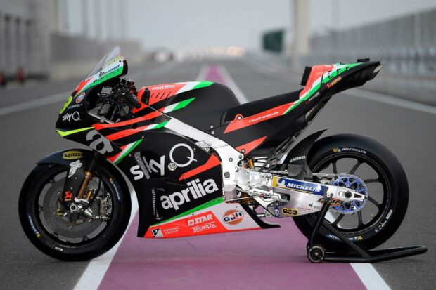 A detailed Aprilia racing motorcycle is parked on a track with sponsor logos visible, HD Desktop Wallpaper