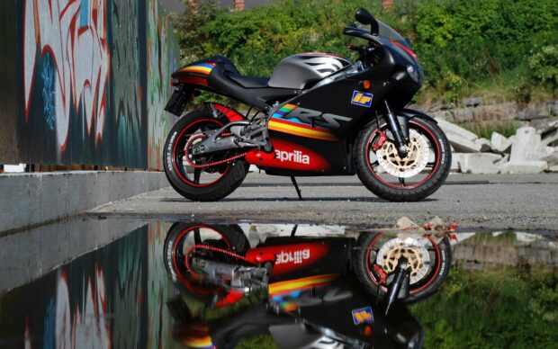 A black Aprilia sports motorcycle is parked beside a graffiti wall with its reflection visible in the puddle below, 2K Desktop Wallpaper