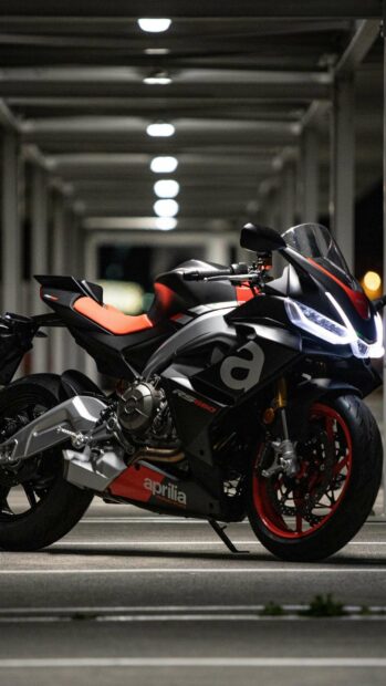 A sleek Aprilia motorcycle is parked in a dimly lit garage with bright headlights, HD Mobile Wallpaper