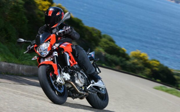 A rider is leaning into a curve on an Aprilia motorcycle along a scenic coastal highway with ocean and vegetation in the background, HD Desktop Wallpaper