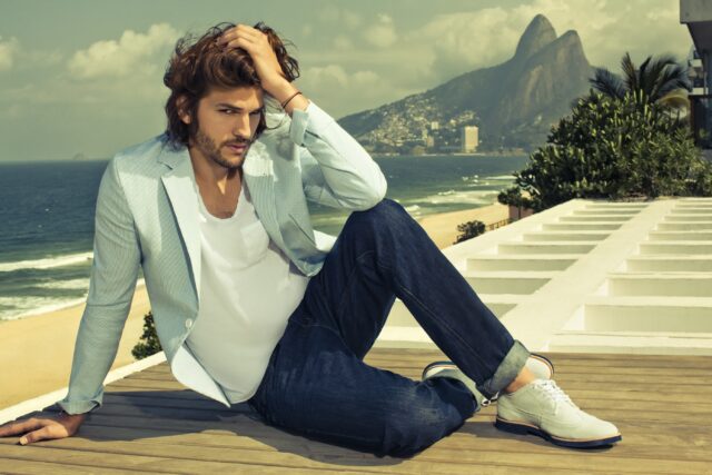 Ashton Kutcher Casual Style Sitting By The Beach With Scenic Mountain Background Wallpapers