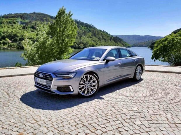 A silver Audi A6 is parked on a stone road by a scenic lake surrounded by green trees and hills, HD Desktop Wallpaper