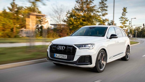 A white Audi Q7 is captured driving swiftly on a city street with blurred trees in the background, showcasing its sleek design, 4K Desktop Wallpaper
