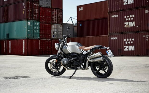 A BMW R Nine T motorcycle is parked on a concrete surface with stacked shipping containers in the background