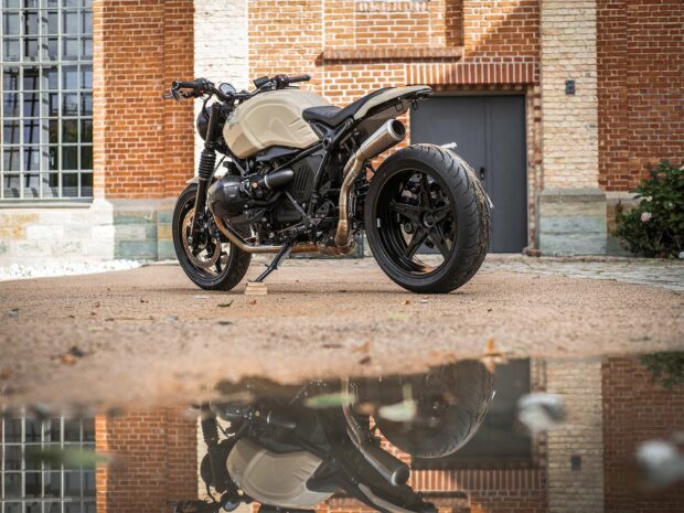 A BMW R Nine T motorcycle is parked outdoors showing its side profile and detailed design with a clear reflection on the wet ground