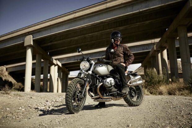 A man wearing a helmet and leather jacket sits on a BMW R Nine T motorcycle on a dirt road beneath a concrete bridge