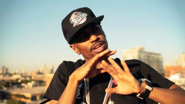 A man wearing a black cap and chains gestures with his hands in front of a city background featuring tall buildings, HD Desktop Wallpaper