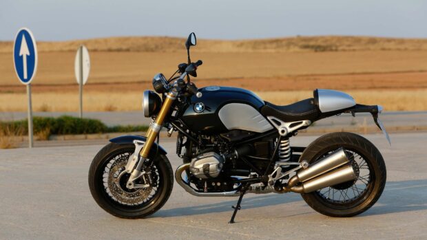 A black BMW R Nine T motorcycle is parked on a desert road with dry grass and blue sky in the background, 2K Desktop Wallpaper