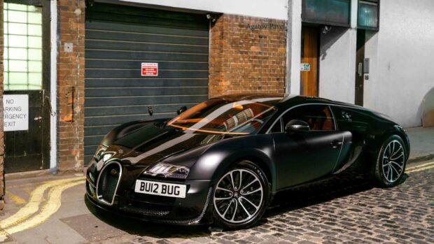 A sleek black Bugatti Veyron is parked on a cobblestone street near a brick wall and garage door, 4K Desktop Wallpaper