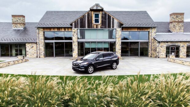 A black Buick Enclave is parked on a stone driveway in front of a large rustic house with stone walls and large windows