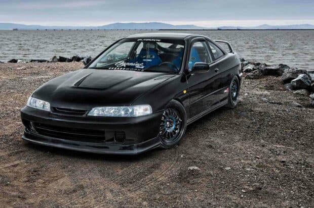 A black Honda Integra is parked on a rocky shore with the ocean and distant mountains in the background, HD Desktop Wallpaper