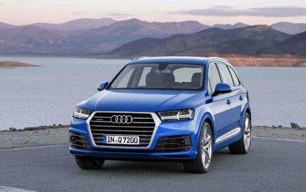 A blue Audi Q7 is parked on a lakeside road with mountains in the background during daylight, 2K Desktop Wallpaper