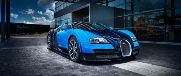 A blue Bugatti Veyron is parked on a paved area in front of a building with large glass windows under a partly cloudy sky, 2K Desktop