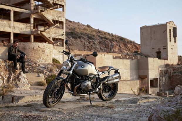 A BMW R Nine T motorcycle is parked on rocky terrain near abandoned buildings with a person sitting in the background, HD Desktop Wallpaper