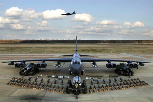 A Boeing B 52 is parked on a runway with an extensive array of weapons displayed in front of it and another B 52 flying overhead