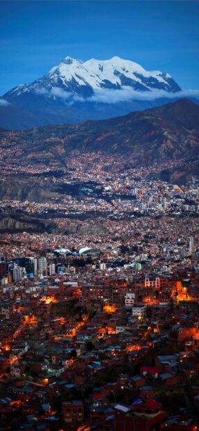 Bolivia Wallpapers show a vibrant cityscape illuminated at dusk with majestic snowy peaks in the background, 2K Mobile Wallpaper